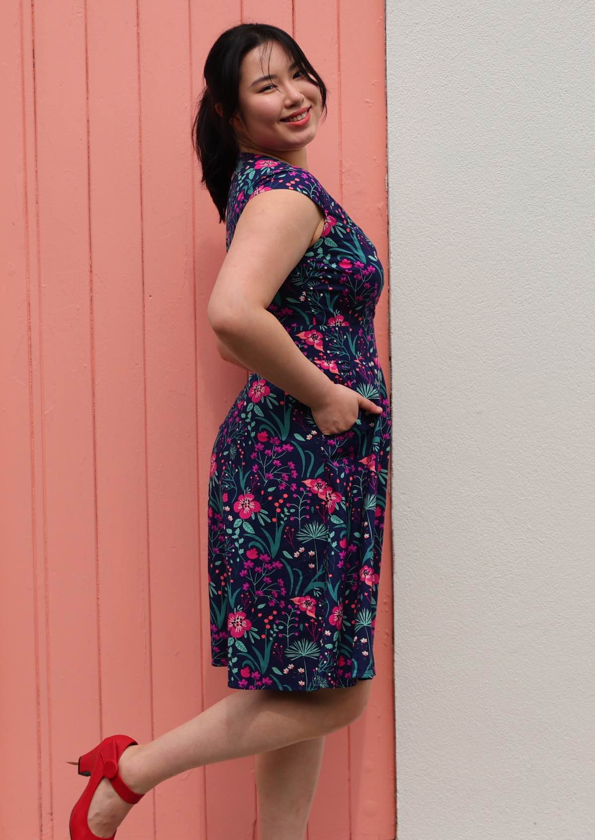 Woman side on n Alice Dress Dianthus retro style 100% cotton navy blue floral with cross over bodice and pockets fit and flare style dress