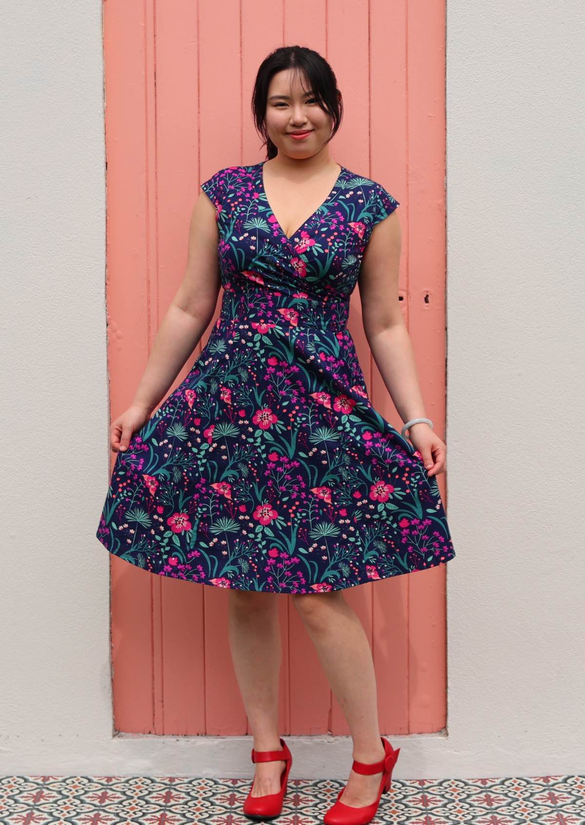 Woman in Alice Dress Dianthus retro style 100% cotton navy blue floral with cross over bodice and pockets fit and flare style dress holding out skirt. Wearing red shoes