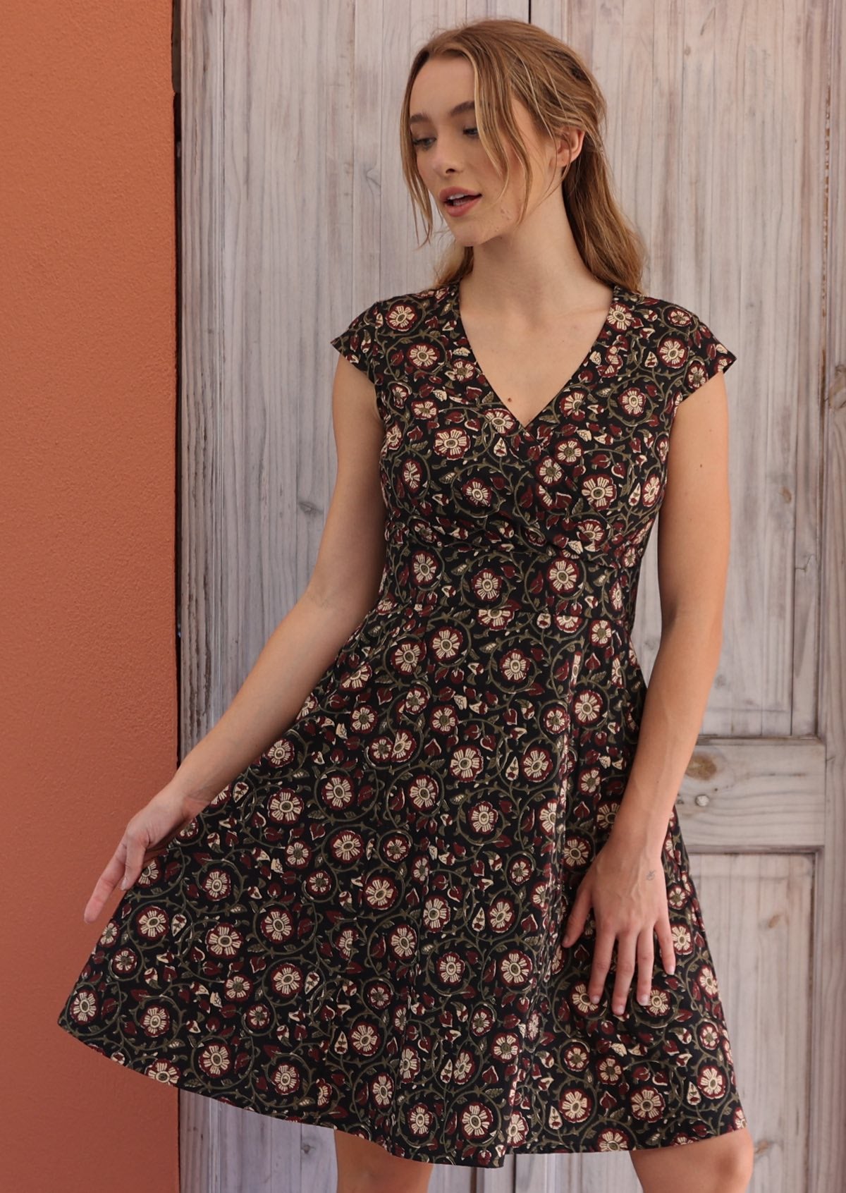 Woman stands in cotton above knee dress with cap sleeves and pockets, in bold maroon floral pattern on black base.