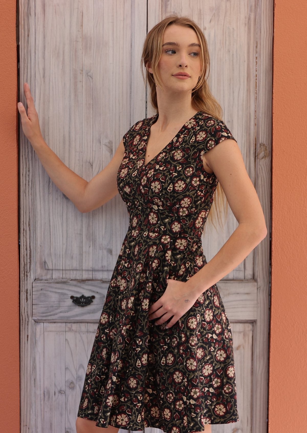 Woman stands sideways in retro style cotton above knee dress with cap sleeves and pockets, in floral print on black base.