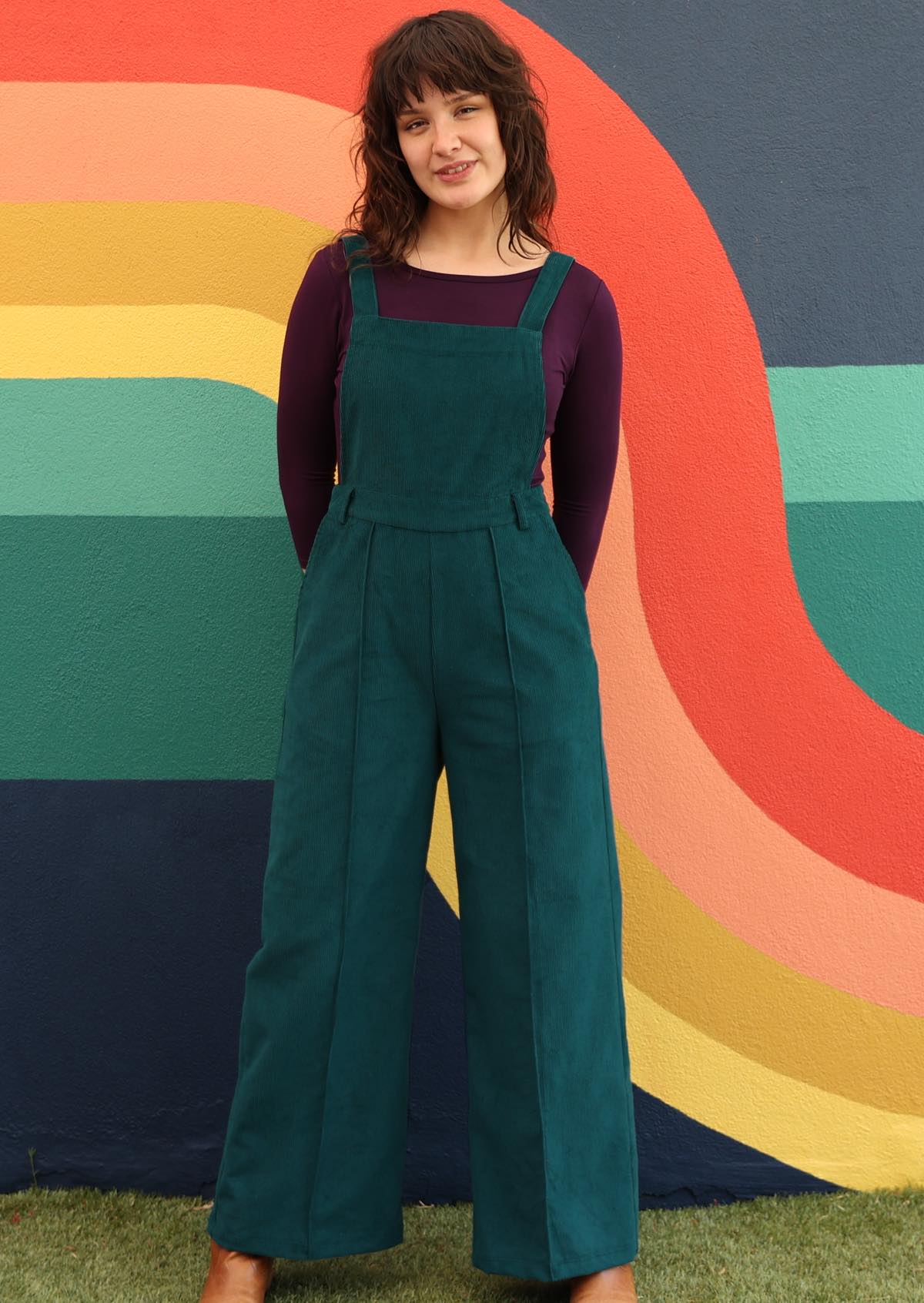 woman wearing dark teal cotton corduroy overalls over long sleeve purple top in front of bright mural 