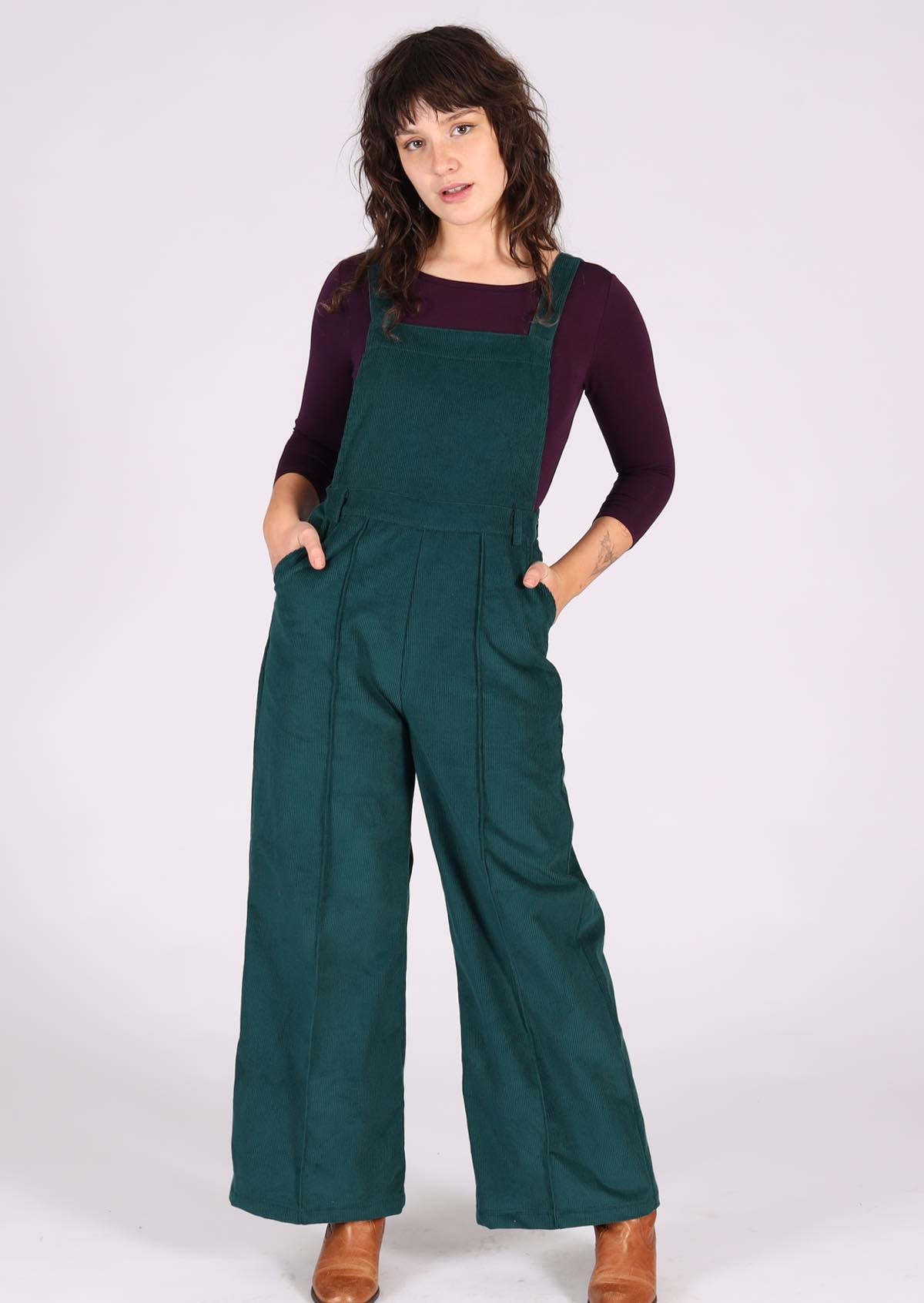 woman wearing dark teal cotton corduroy overalls over long sleeve purple top with hands in pockets