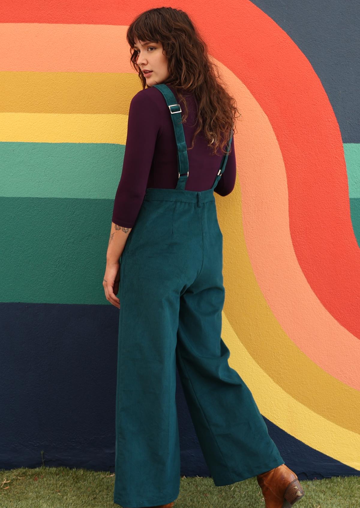 woman wearing dark teal cotton corduroy overalls over long sleeve purple top in front of bright mural  back view