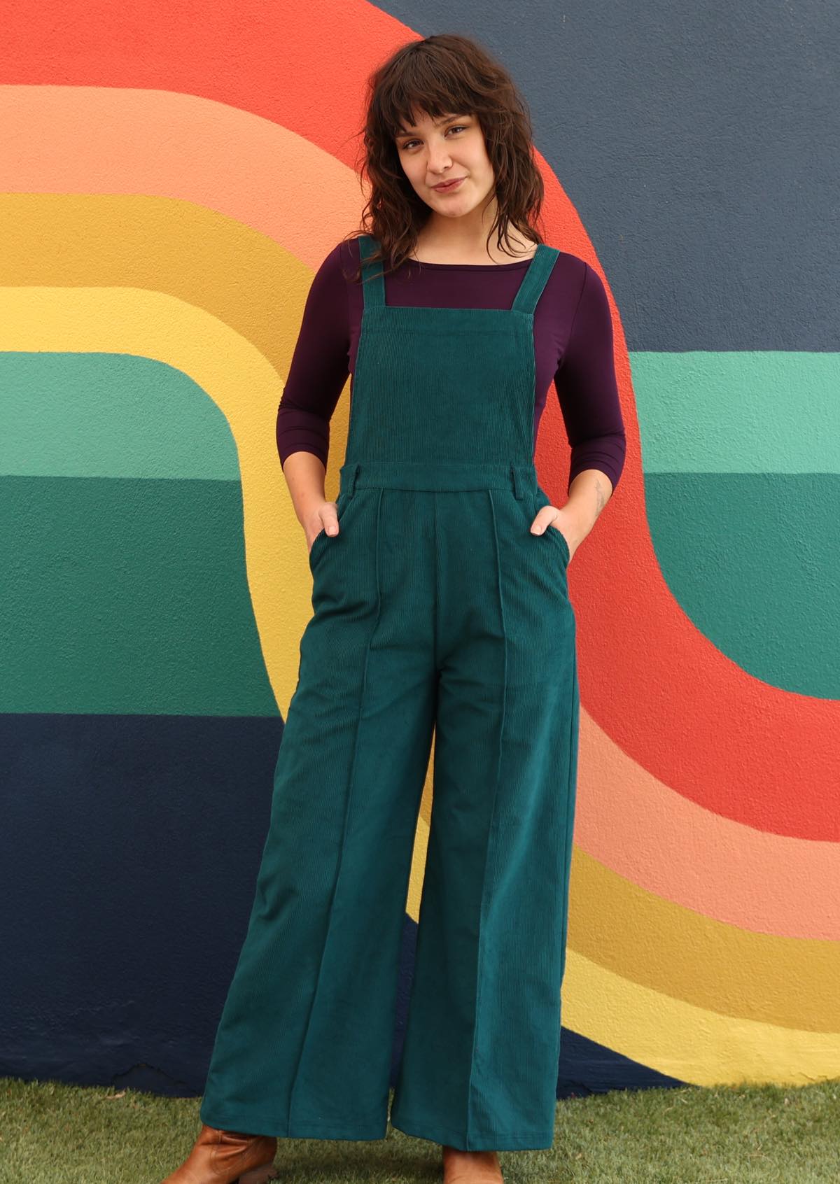 woman wearing dark teal cotton corduroy overalls over long sleeve purple top in front of bright mural wall
