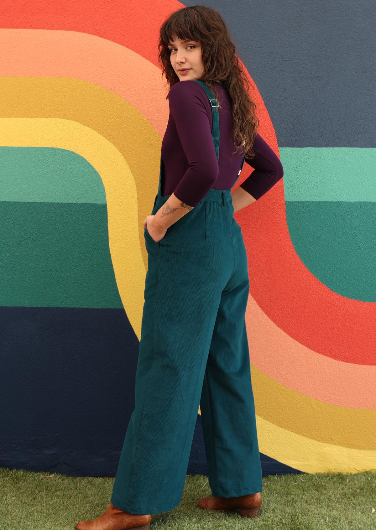 woman wearing dark teal cotton corduroy overalls over long sleeve purple top in front of bright mural side view