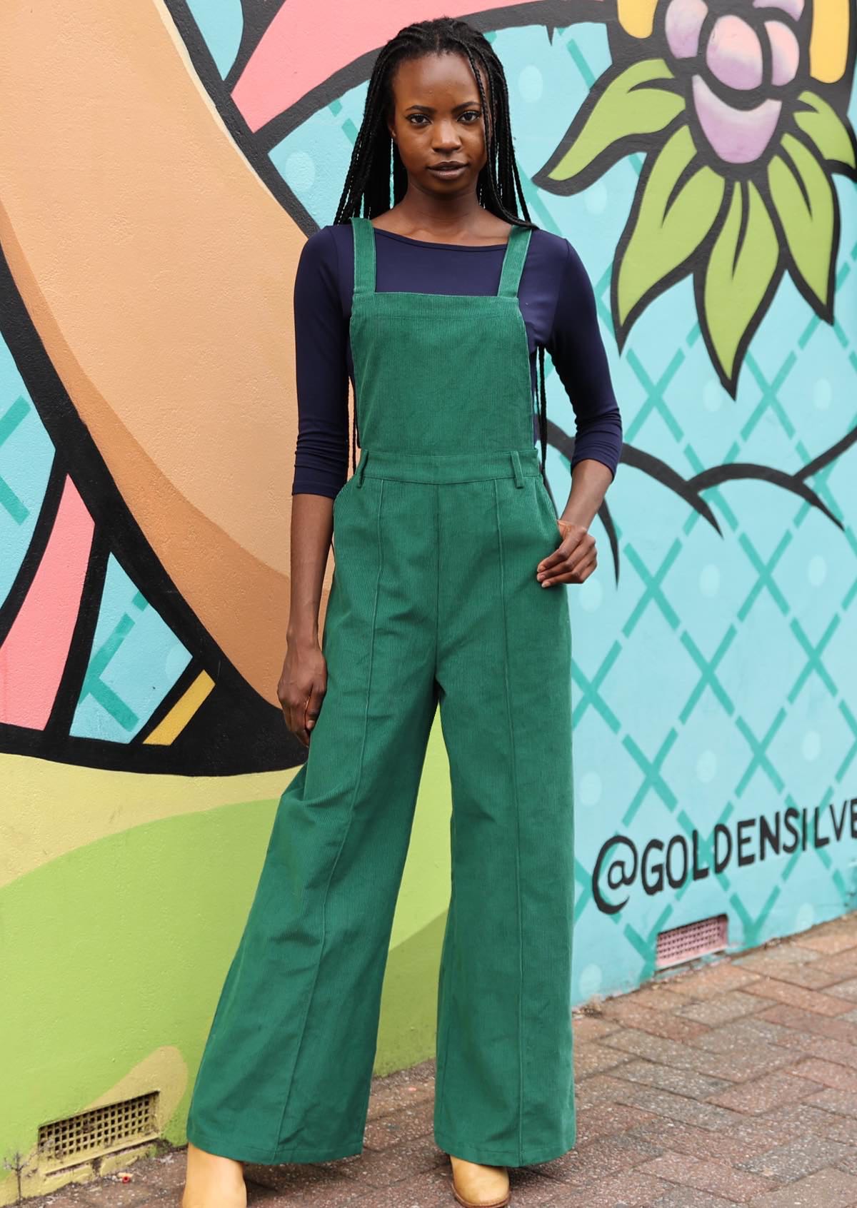 woman wearing green cotton corduroy overalls with hands in pockets in front of mural