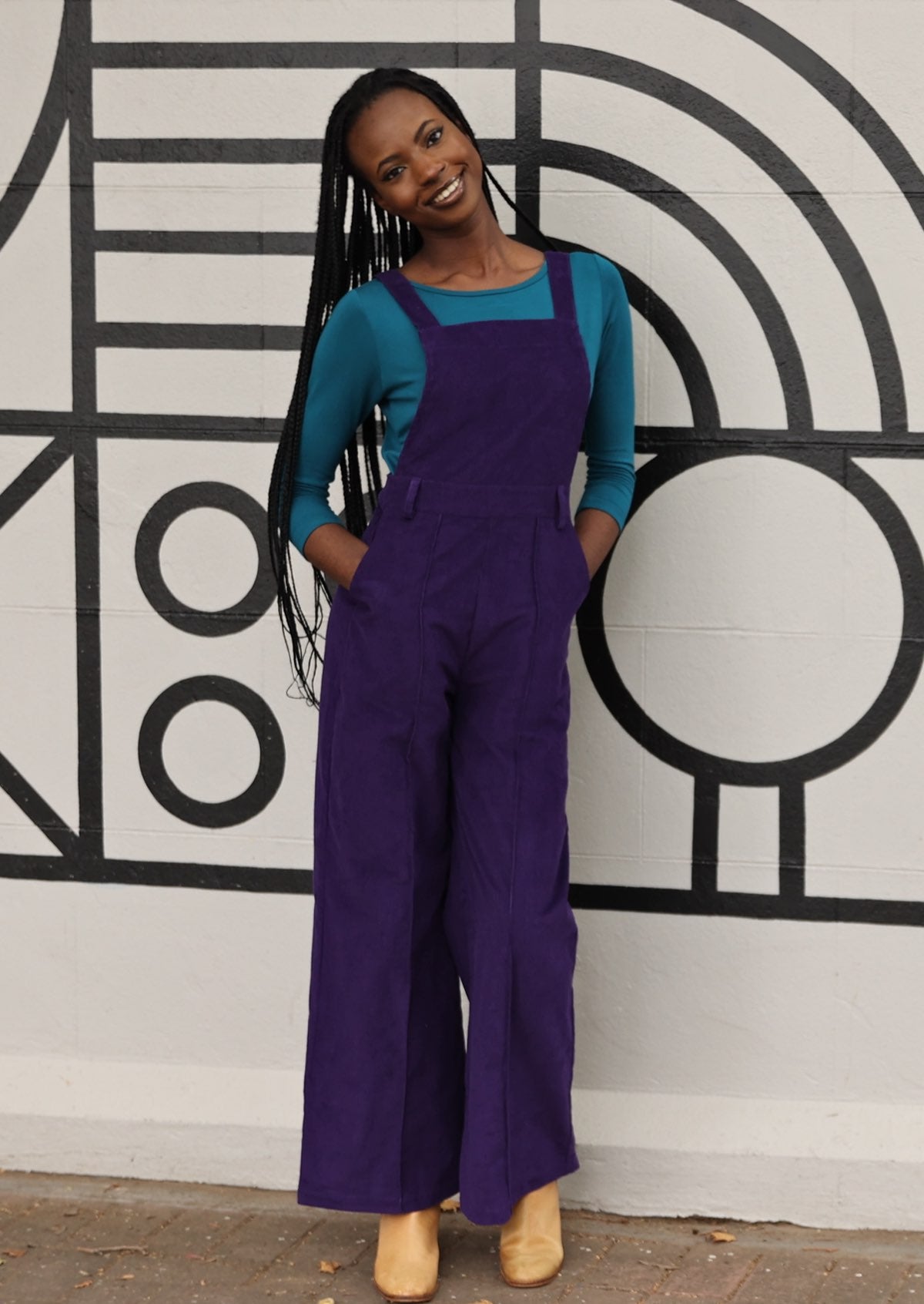 model wearing purple cotton corduroy overalls with hands in pockets in front of mural