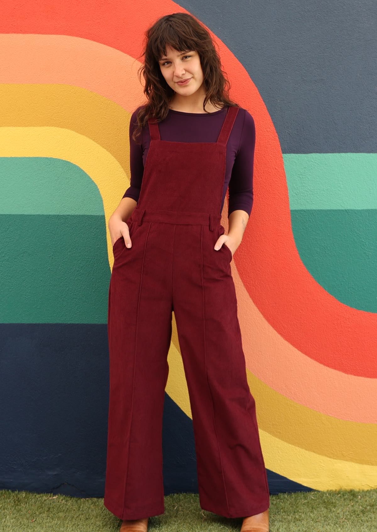 woman wearing maroon cotton corduroy overalls over long sleeved purple top, with hands in pockets