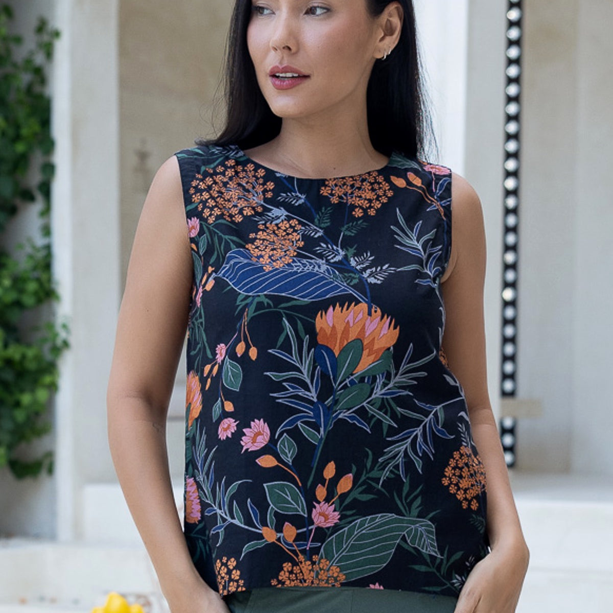 Model standing with hands in pocket of pants, wearing a sleeveless cotton top with round neckline and A line cut in beautiful bold botanical print on black base
