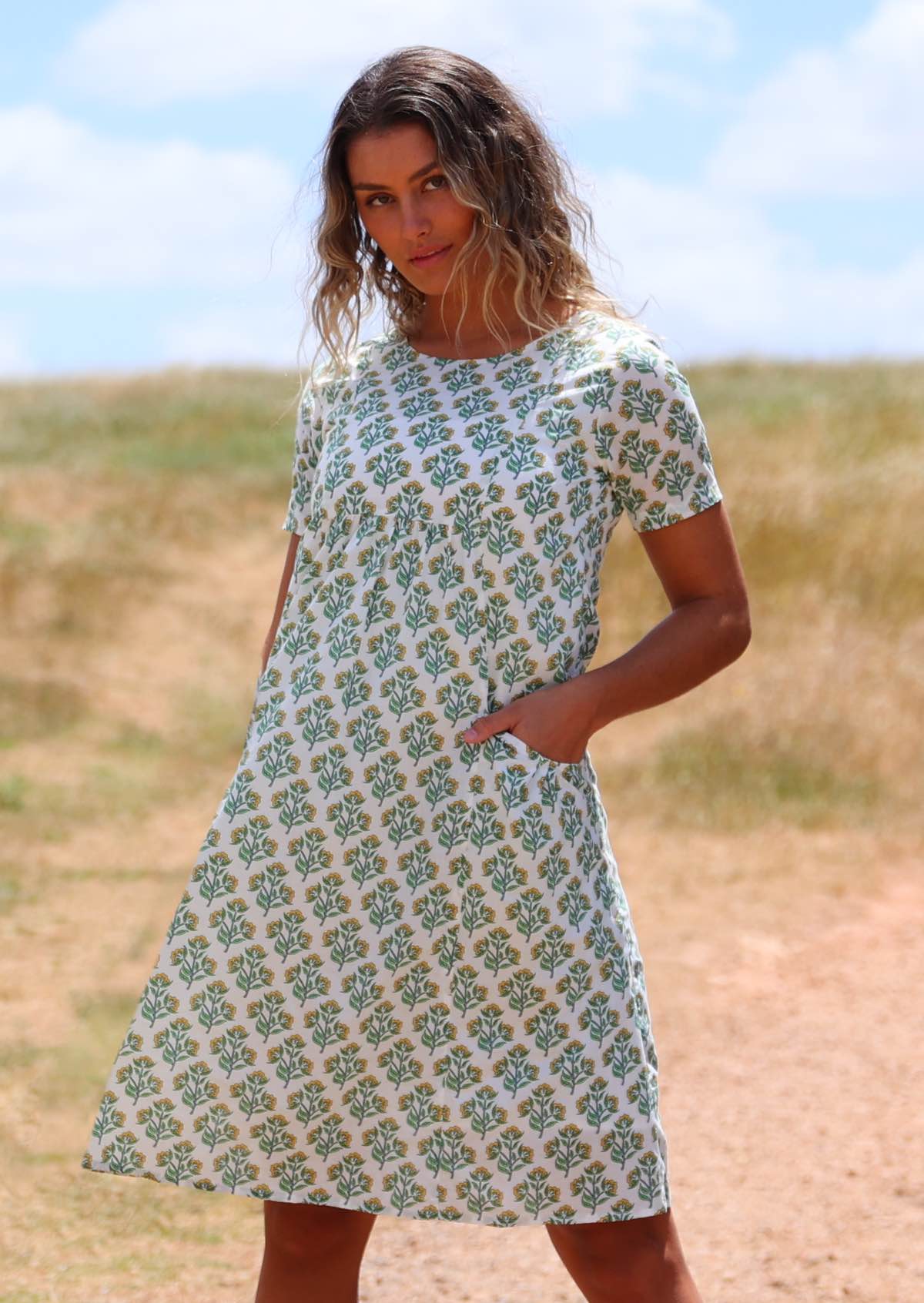 Woman wearing white cotton sundress with floral pattern on grassy hill with hands in pockets