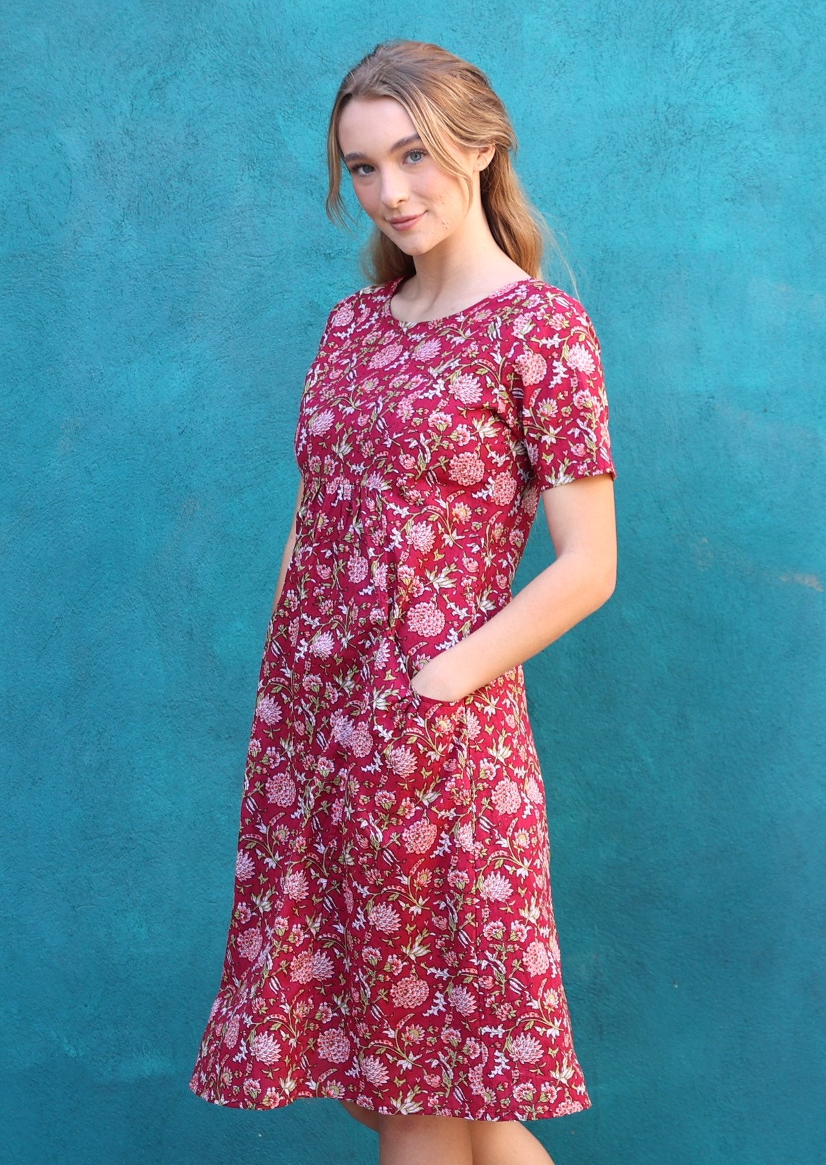 Woman wearing cranberry based floral print cotton dress with an empire waistline and her hands in the deep pockets