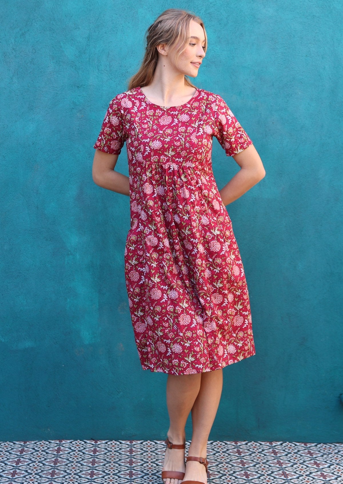 Woman stands in short sleeve sun dress with floral print on a cranberry base with hands behind back