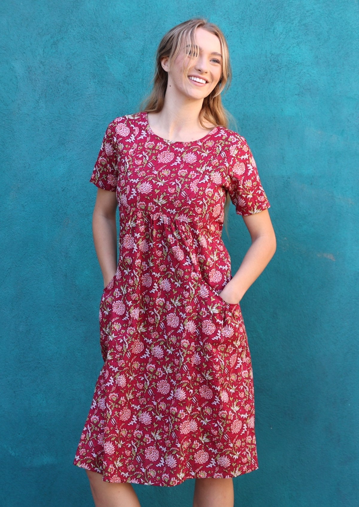 Model poses in cotton short sleeve sun dress with round neckline and small gathers under the bust with her hands in the deep pockets