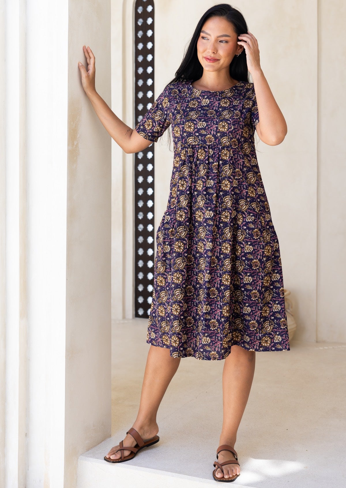 Woman wearing a purple floral cotton dress standing against a neutral wall.