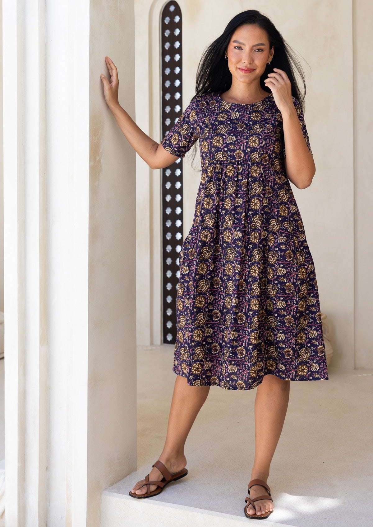Woman wearing a purple floral sun dress standing against a white wall.