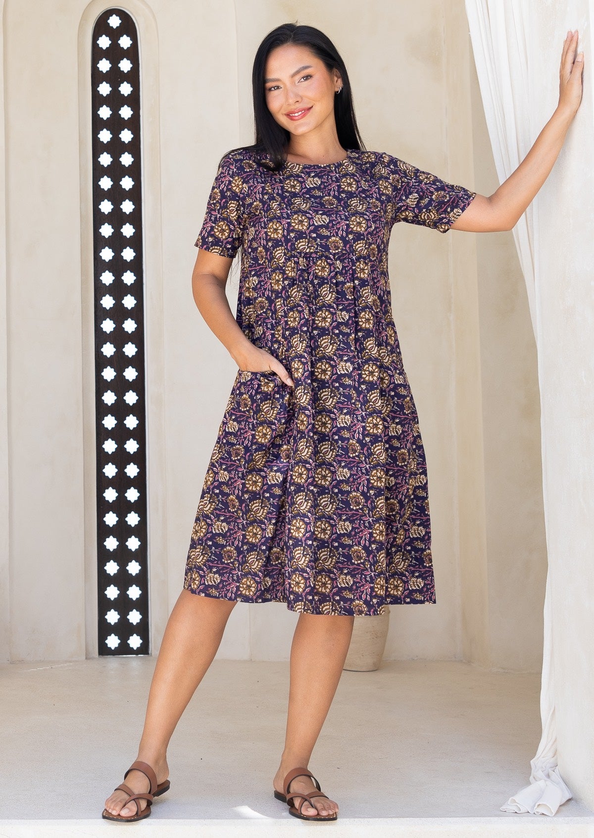 Woman wearing a floral dress standing in a bright room with white walls and a decorative wall panel.