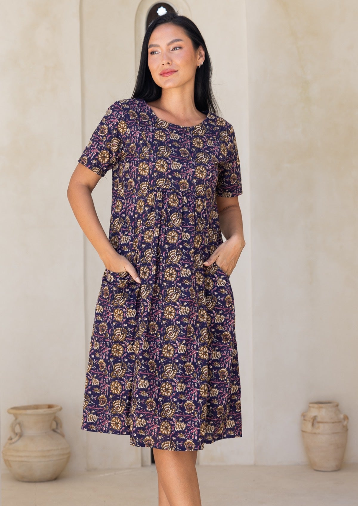 Woman wearing a purple floral dress with hands iin pockets, standing against a plain wall.