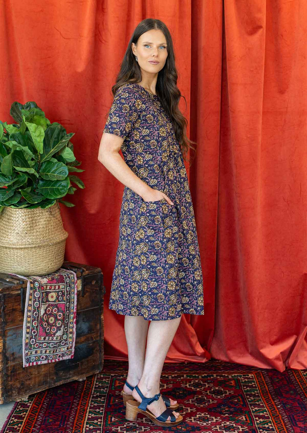 Woman stands in short sleeve sun dress with pink and cream floral print on a deep purple base with hand in pocket.