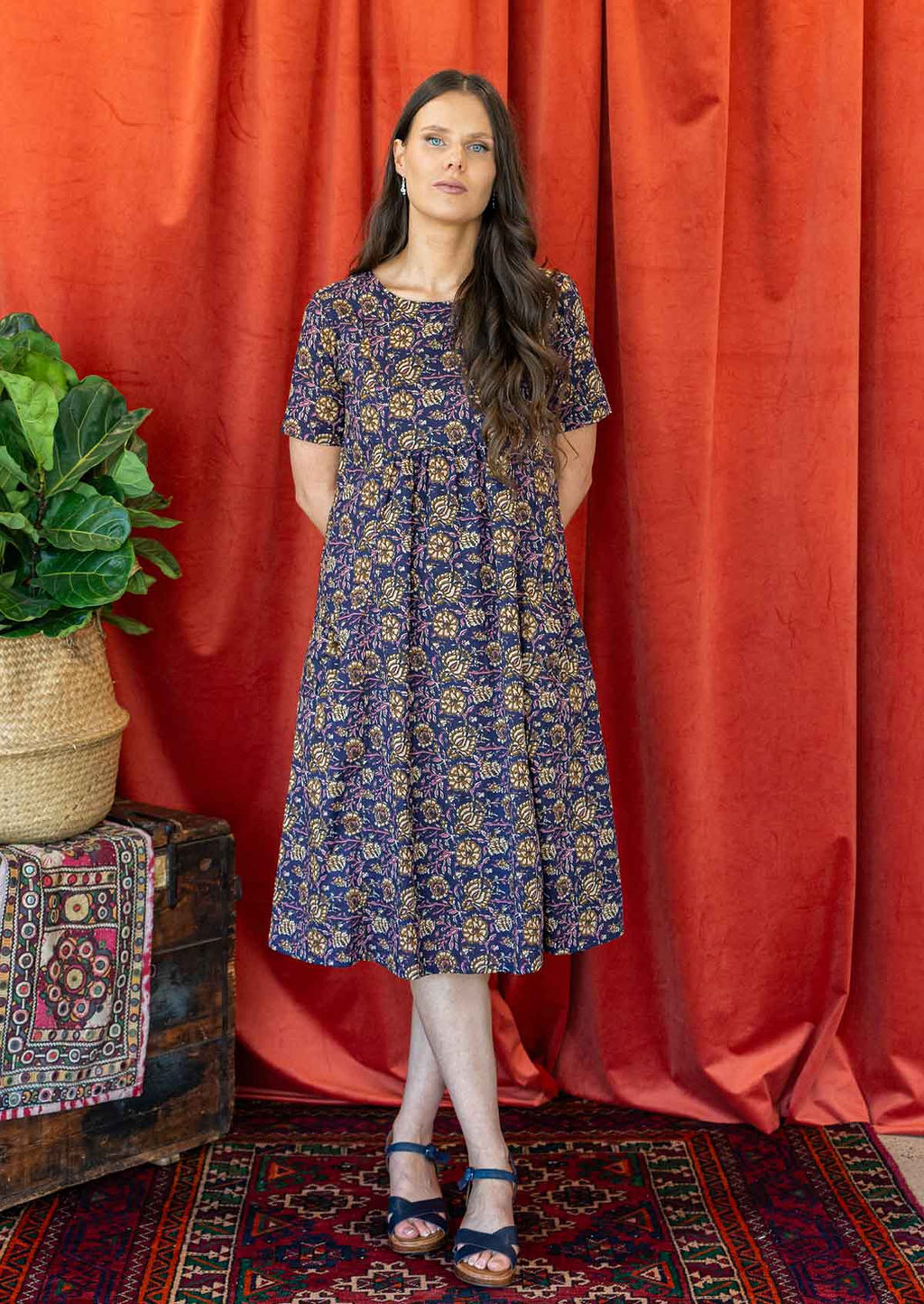 Woman with her hands back wears a beautiful floral print dark purple dress with short sleeves, round neckline and gathers under the bust.