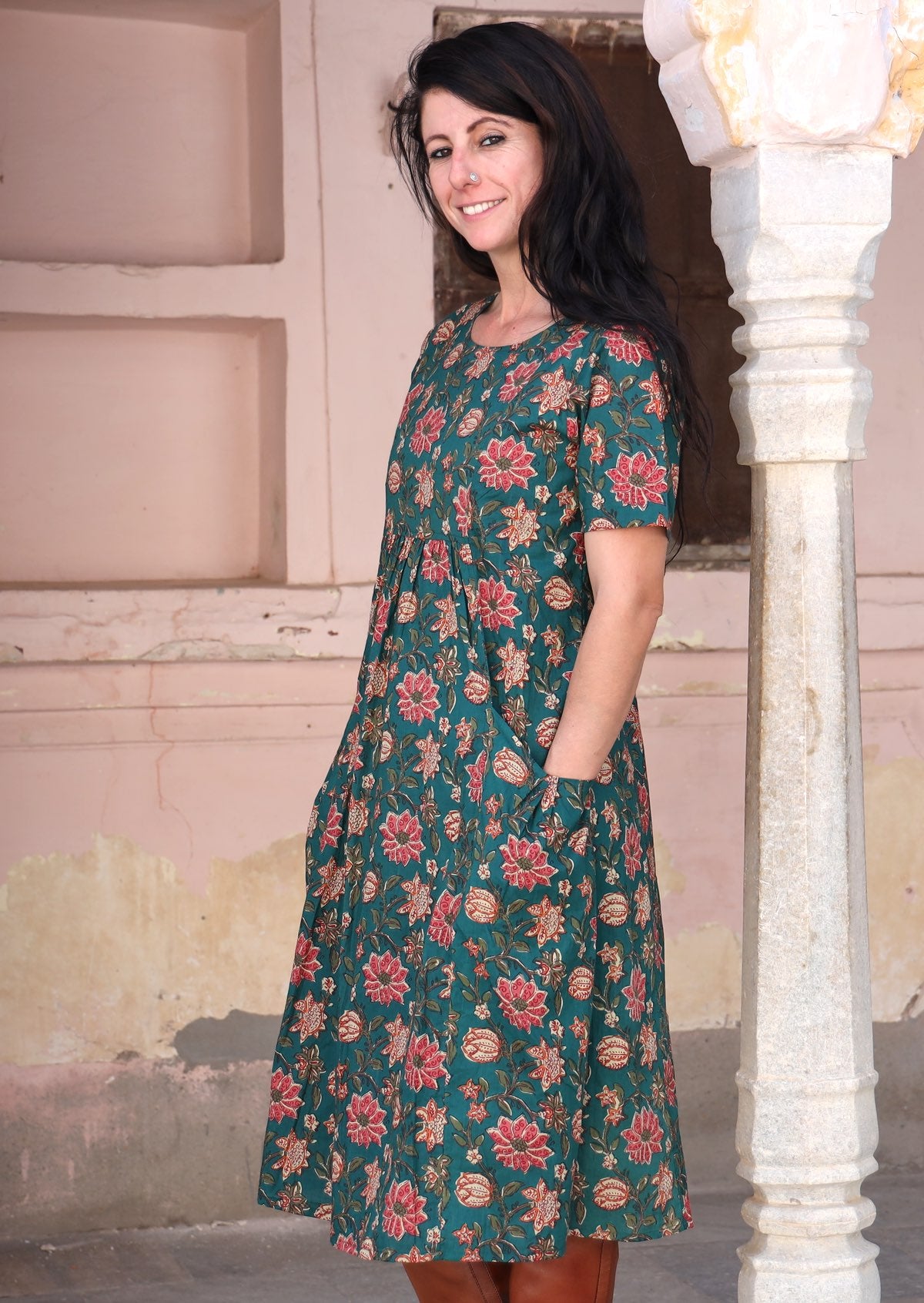 Model stands sideways in cotton knee length dress with burgundy floral print on dark green base.