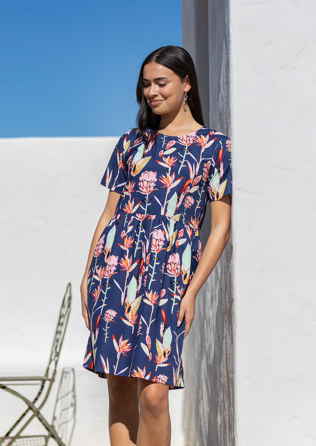 Woman standing against the white wall in the sun wears relaxed fit above knee length dress with short sleeves and pockets.