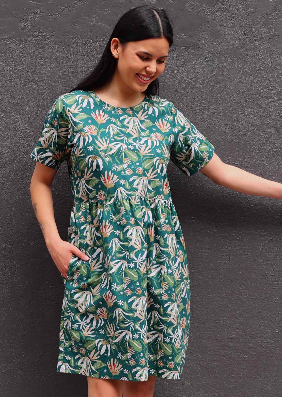 Model with dark hair in loose fit green cotton floral dress with pockets in front of dark grey wall