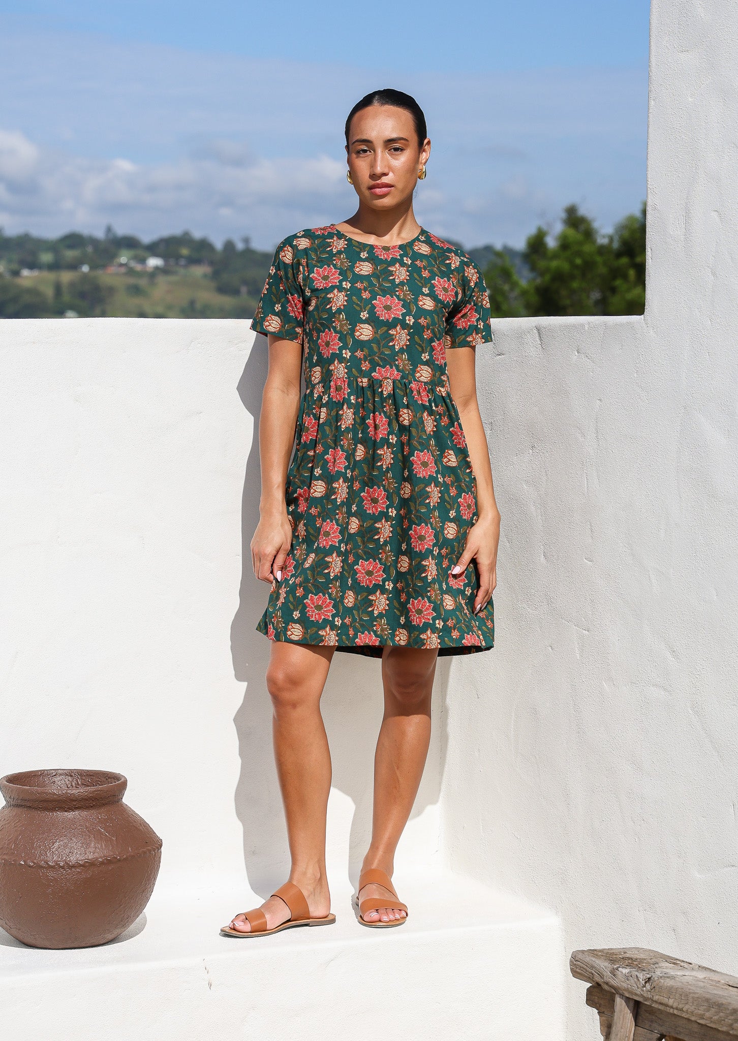 Woman wearing relaxed fit, above the knee length cotton dress with short sleeves and bright floral print on deep green base.
