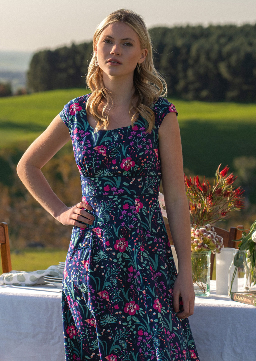 woman in retro inspired Scarlett Dress Dianthus in a vibrant navy, purple, pink and aqua floral print on 100% cotton in front of laid out table