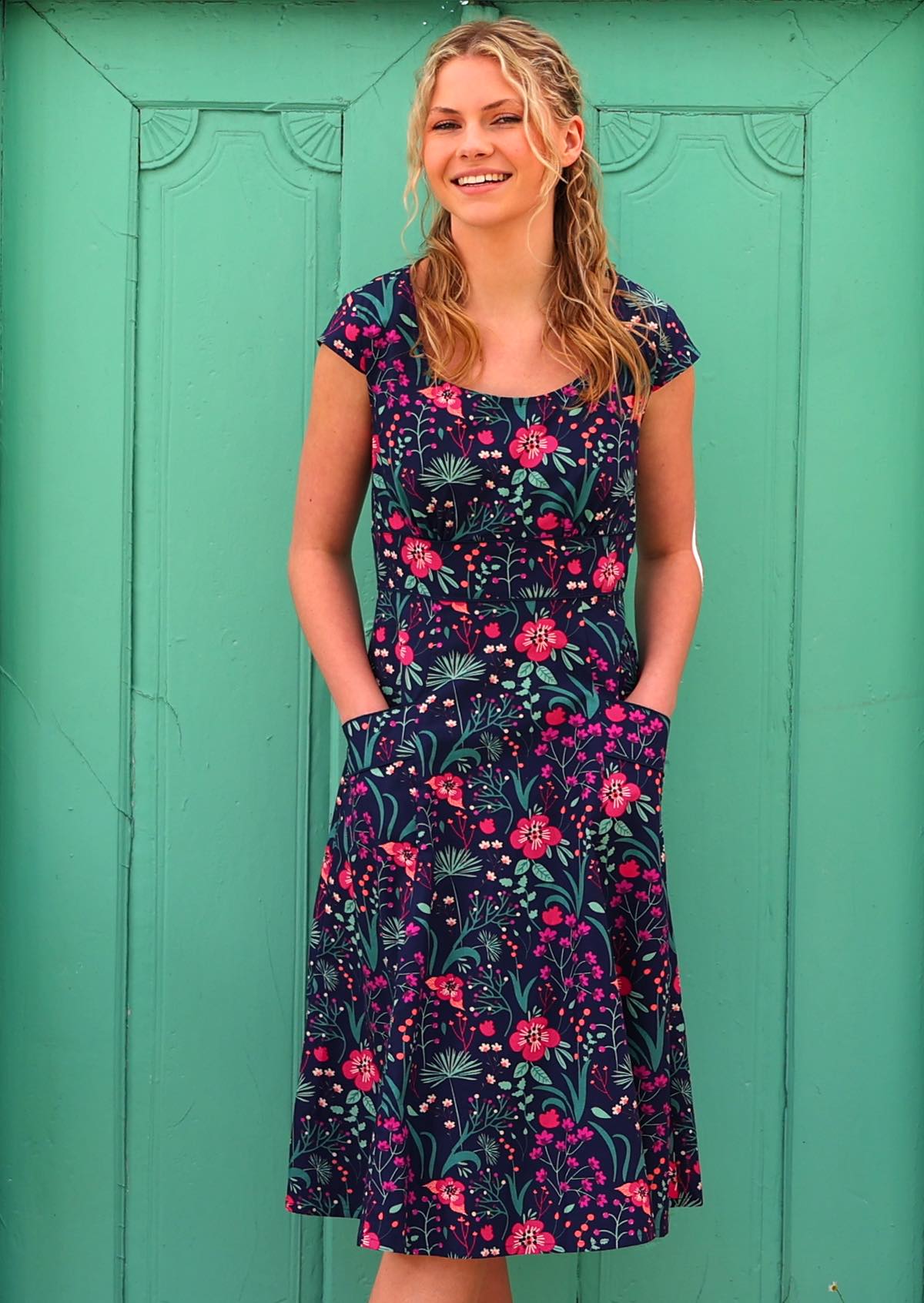 woman in front of aqua door in retro inspired Scarlett Dress Dianthus vibrant navy, purple, pink and aqua floral print on 100% cotton