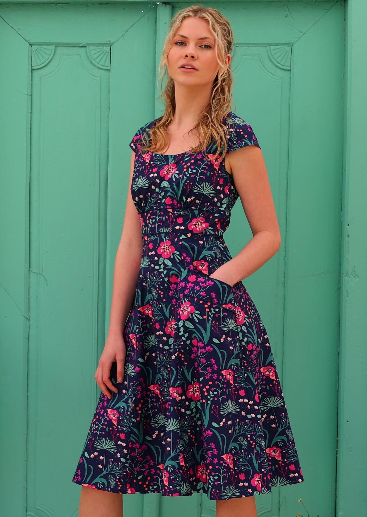 woman in retro inspired Scarlett Dress Dianthus in a vibrant navy, purple, pink and aqua floral print on 100% cotton with hand in pocket in front of aqua door