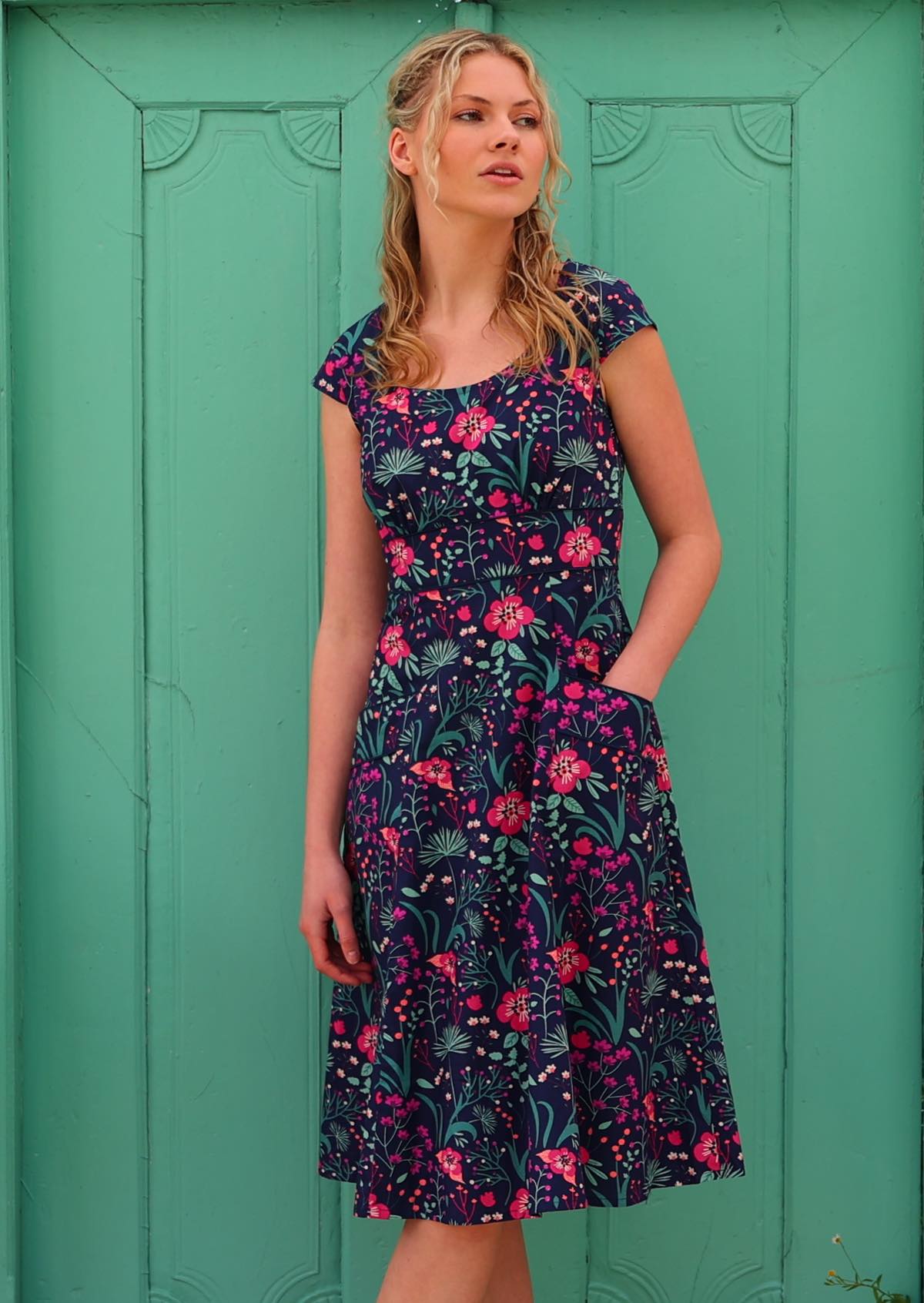 woman in retro inspired Scarlett Dress Dianthus in a vibrant navy, purple, pink and aqua floral print on 100% cotton with hand in pocket in front aqua door