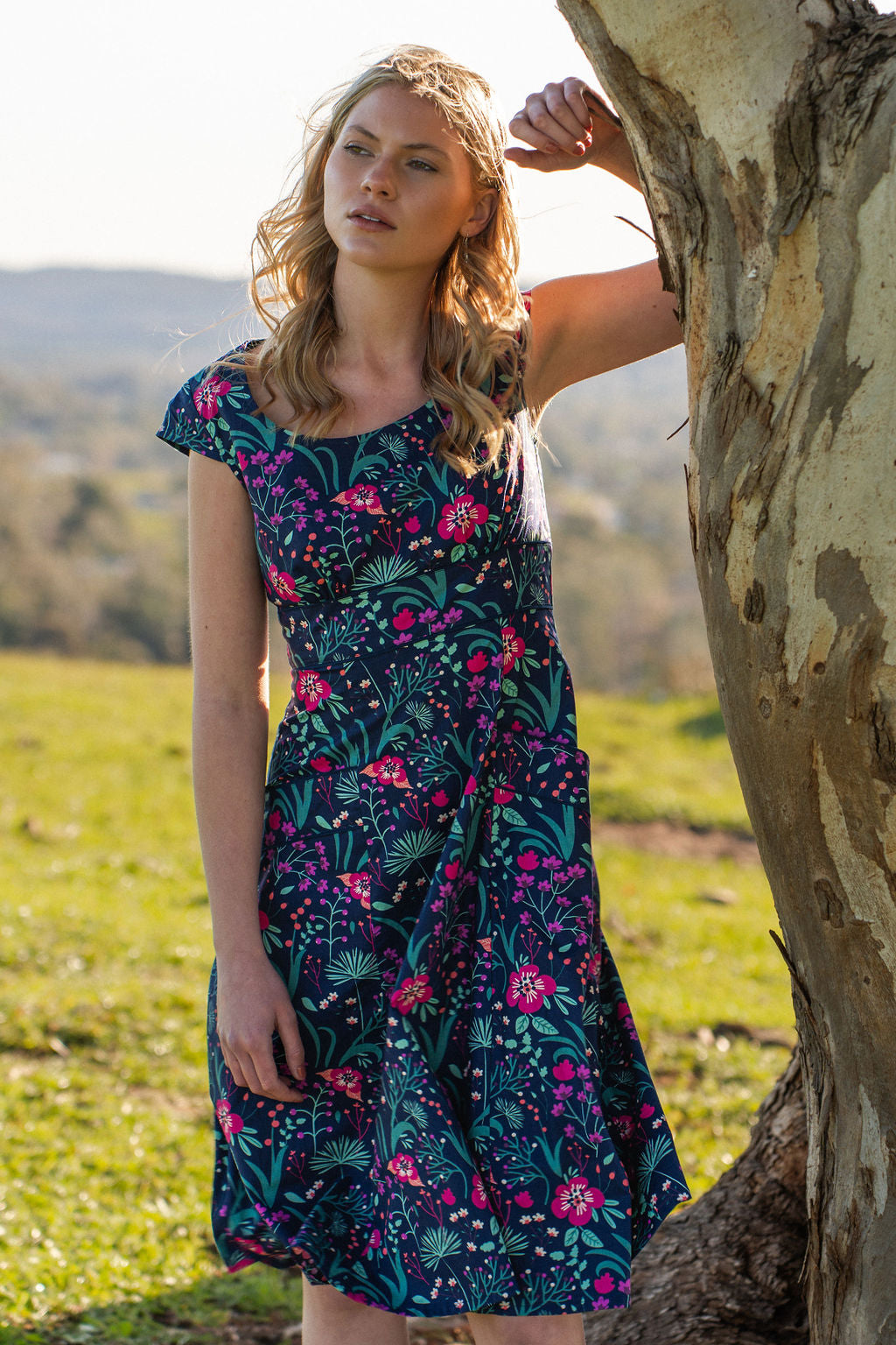woman in retro inspired Scarlett Dress Dianthus in a vibrant navy, purple, pink and aqua floral print on 100% cotton under gum tree with arm up