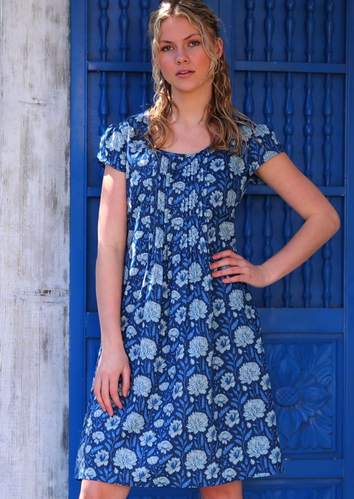 Woman with blonde hair wearing a blue cotton dress with cap sleeves and pin tucks across bodice in light blue and white floral design, in front of blue door