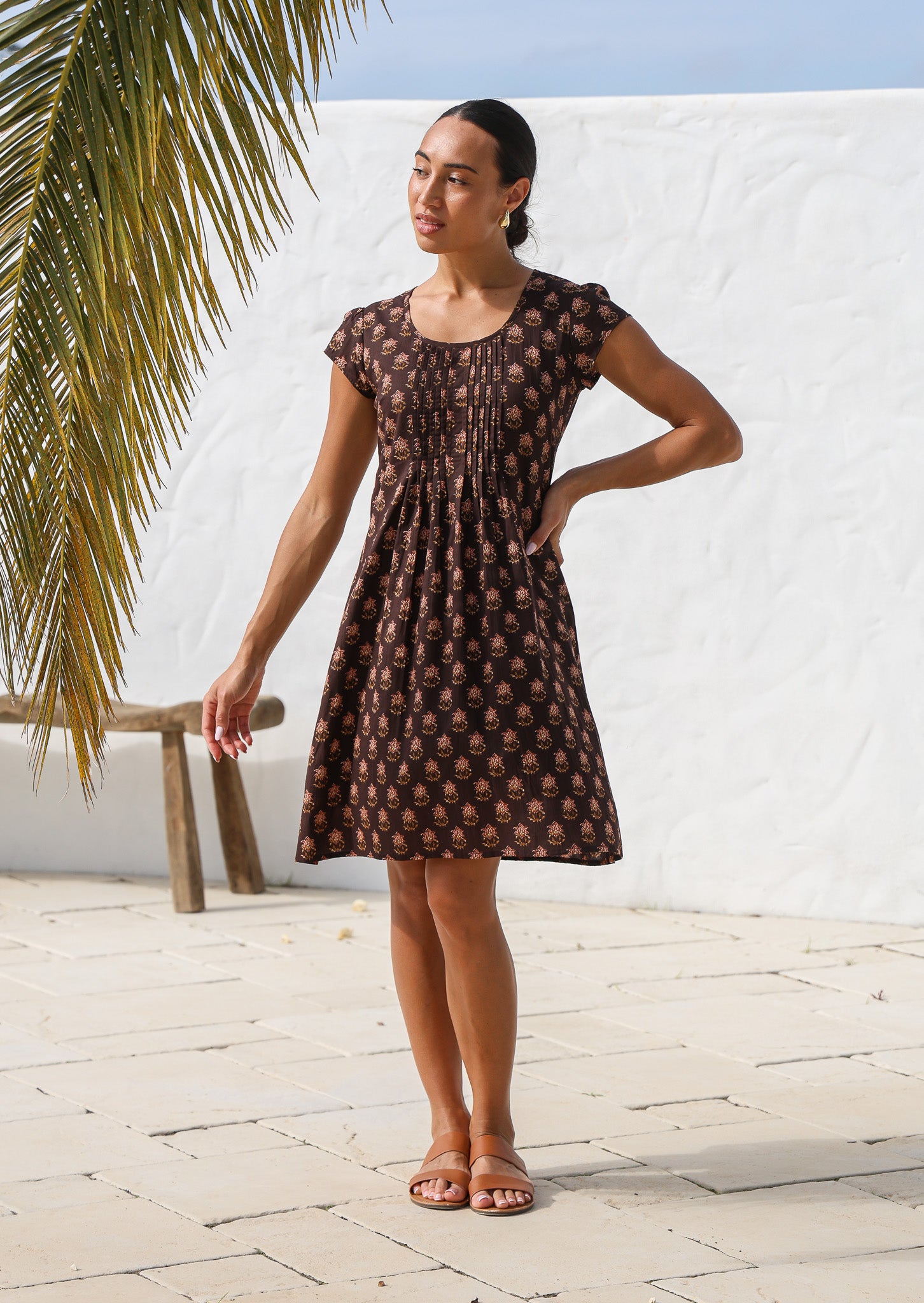 Women standing in sun wears a cotton mini dress with caped sleeves, round neckline and pleated bodice.