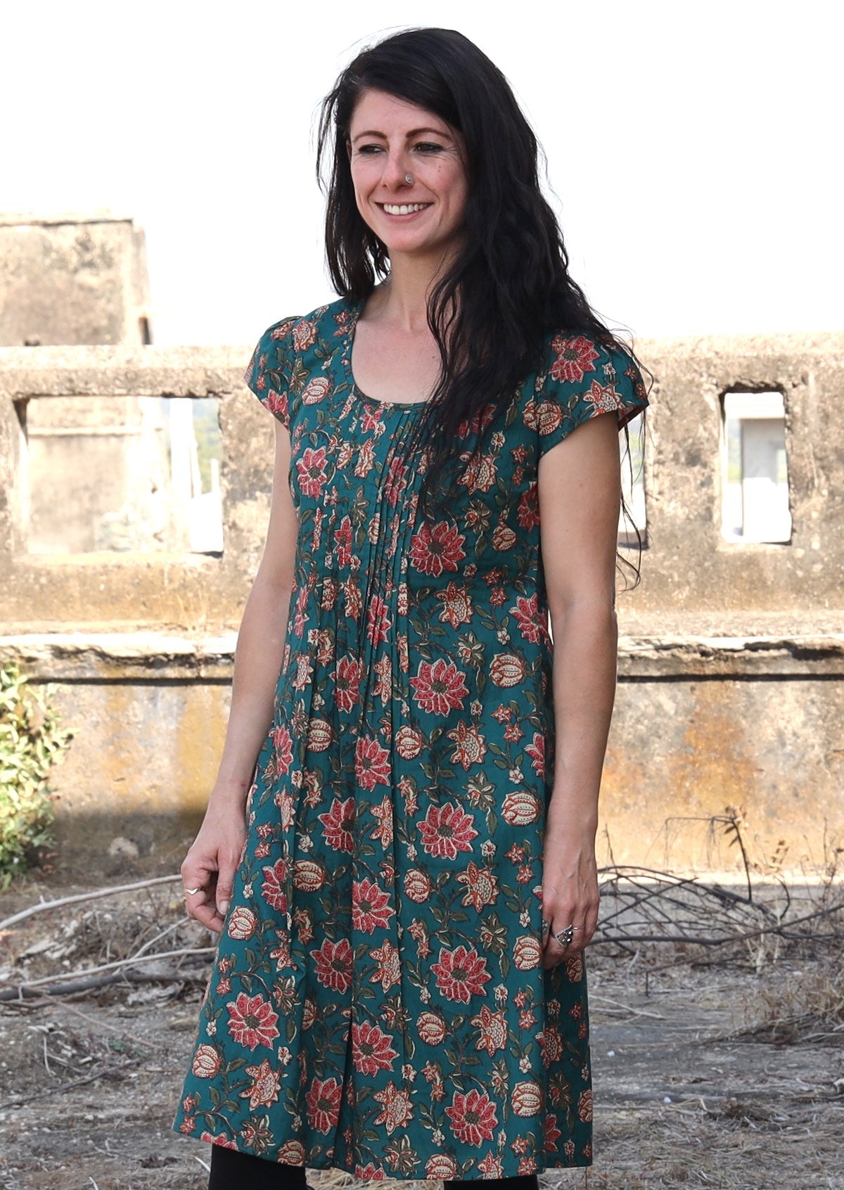 Women standing slightly sideways wears a green based cotton dress with short sleeves, round neck an pleated bodice.