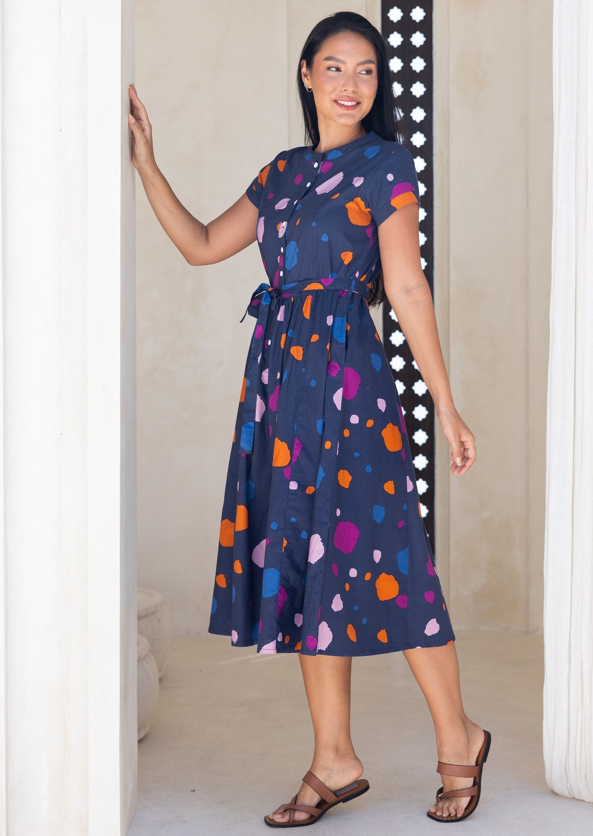Woman stands side on in a navy based cotton 50's style dress with colourful polka dot print, with cap sleeves and a waist tie