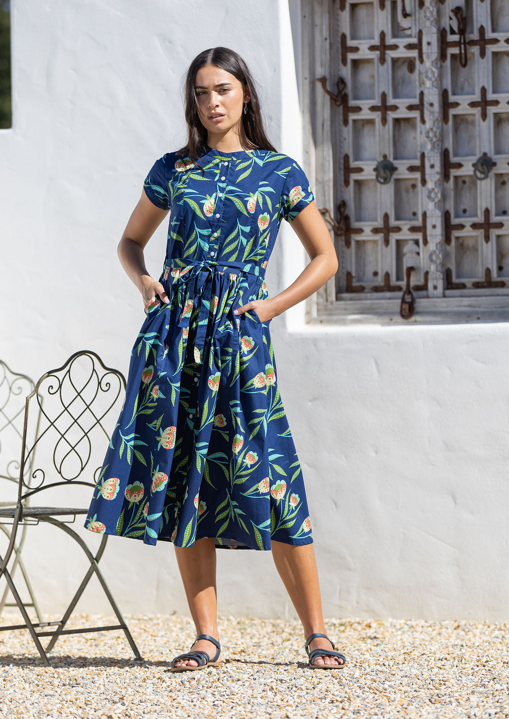 Woman wearing a blue floral cotton dress standing in front of a white wall with hands in her pockets
