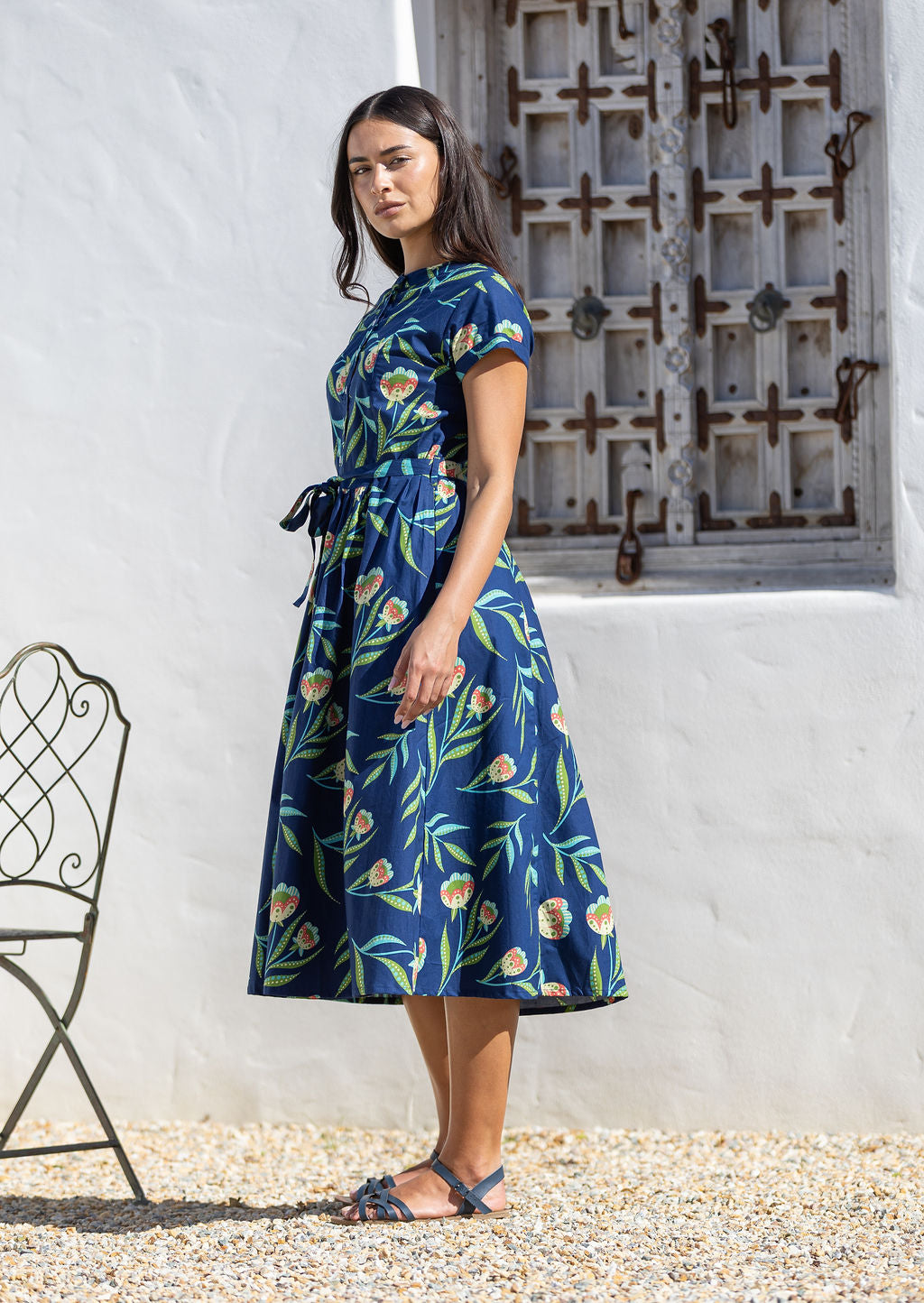 Woman wearing a blue cotton floral dress with full skirt standing in front of a white wall with a decorative metal gate.