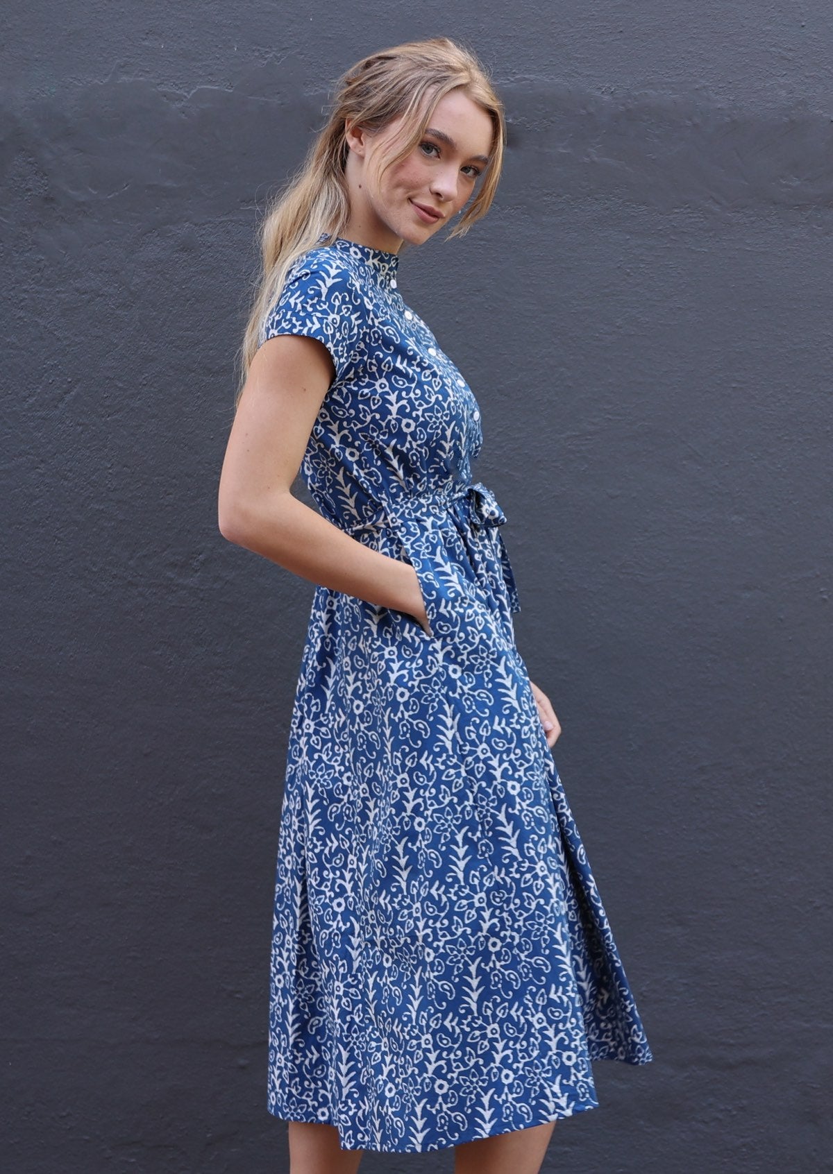 Woman poses in cotton midi length dress in blue based floral print with her hand in hidden side pocket