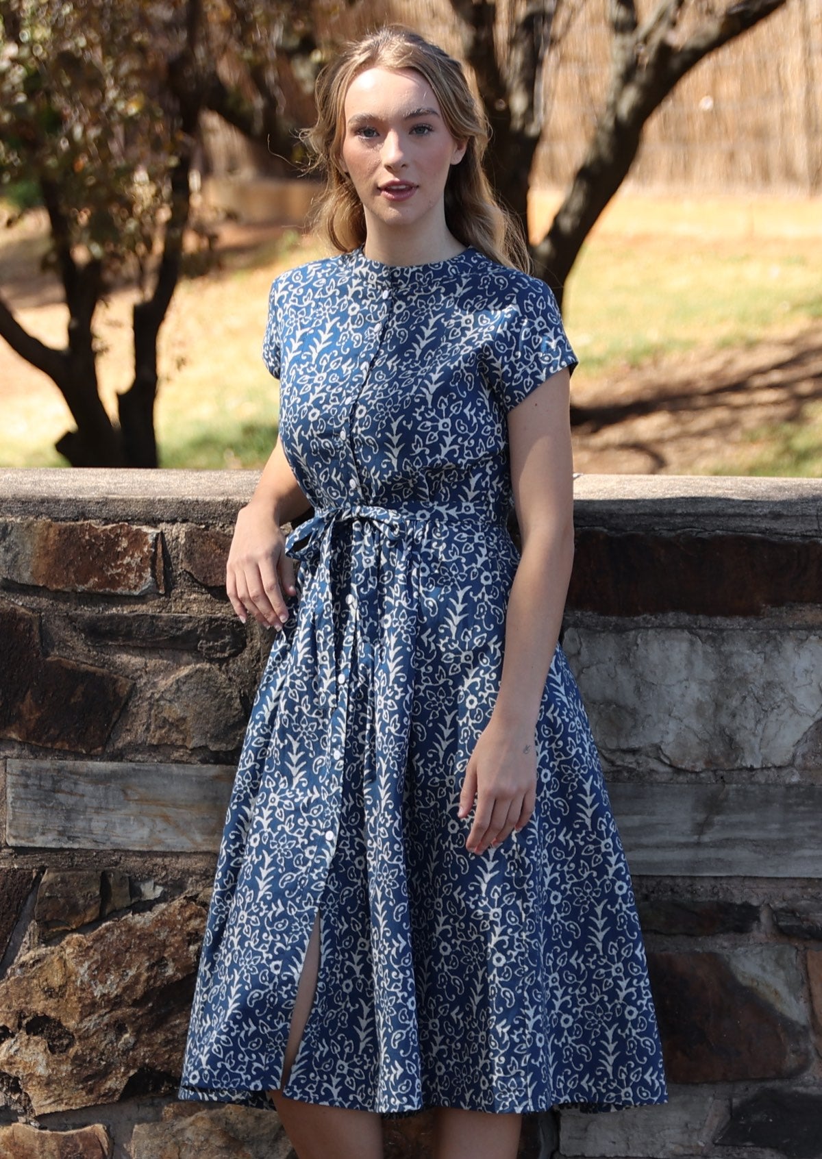 Woman leans on stone wall in blue based floral print cotton button through 50's style dress