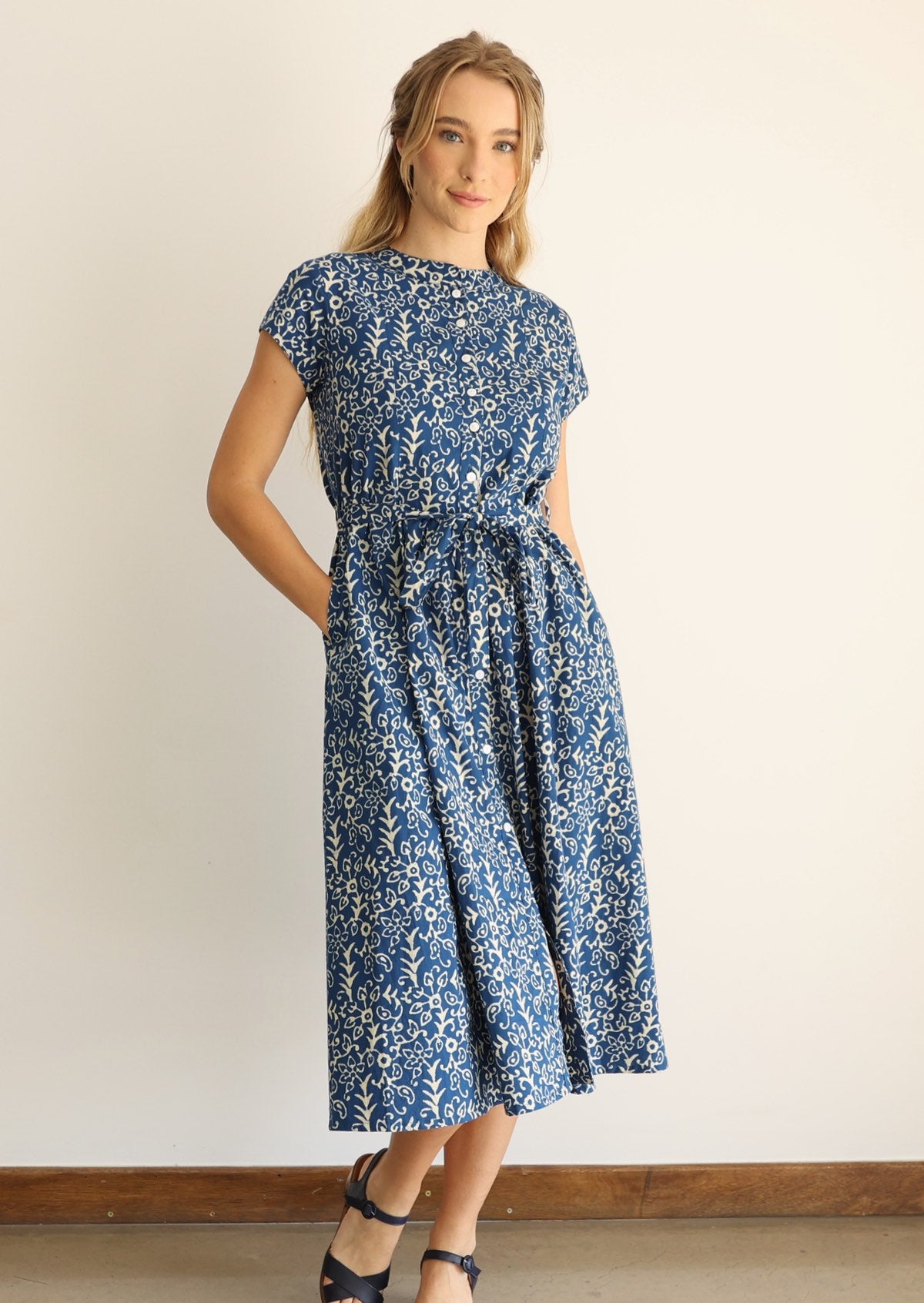 Woman wears cotton button through 50's style dress in floral print on a blue base, with hands in hidden side pockets