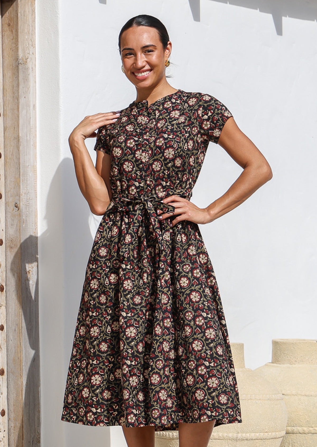 Woman wears black based floral printed cotton button through dress with hidden side pockets, short sleeves and a waist tie.