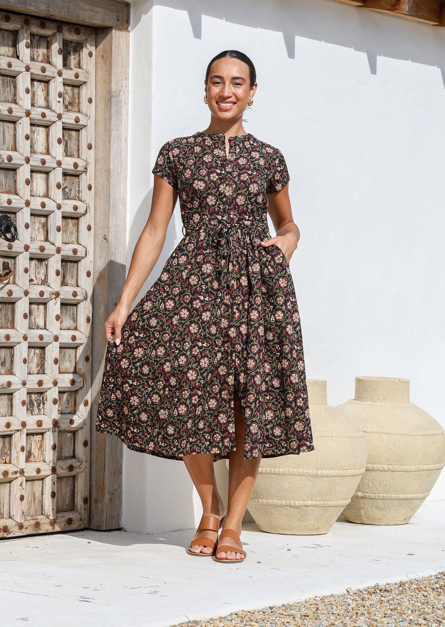 Woman wears 50's style cotton button through midi length dress with high round neckline, short sleeves and a waist tie.