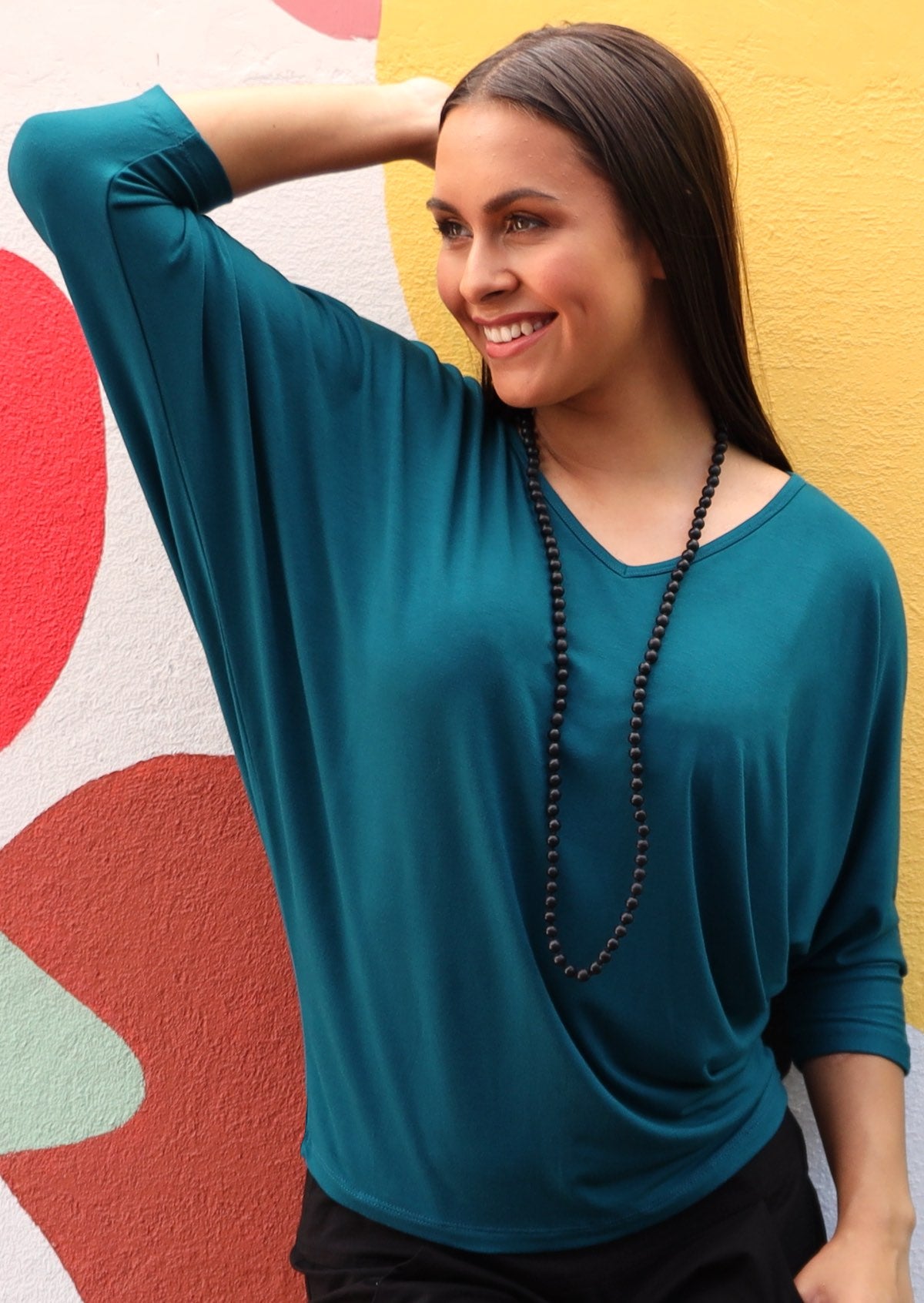 Woman wearing a 3/4 sleeve rayon batwing v-neck teal top in front of colourful wall.