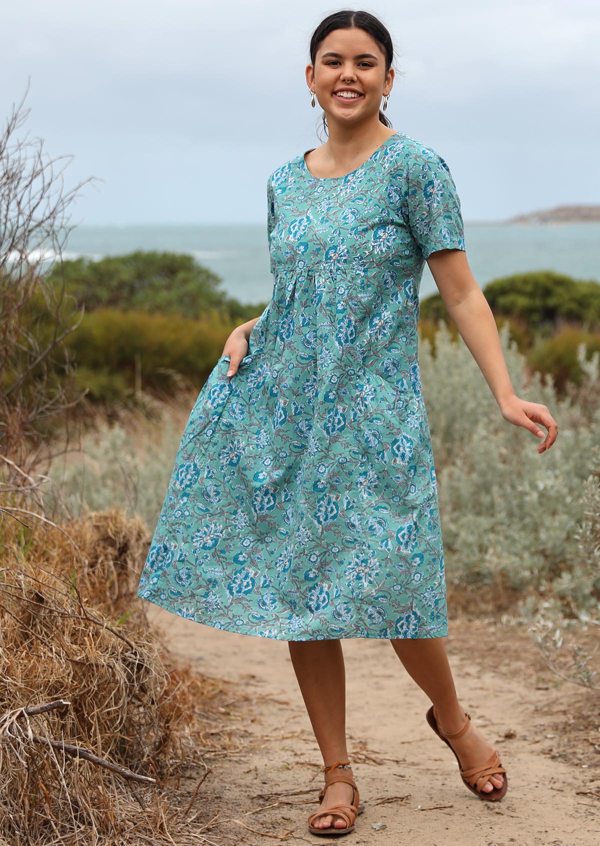 Model posing in knee length, 100% cotton dress in Indian floral print on aqua blue base.