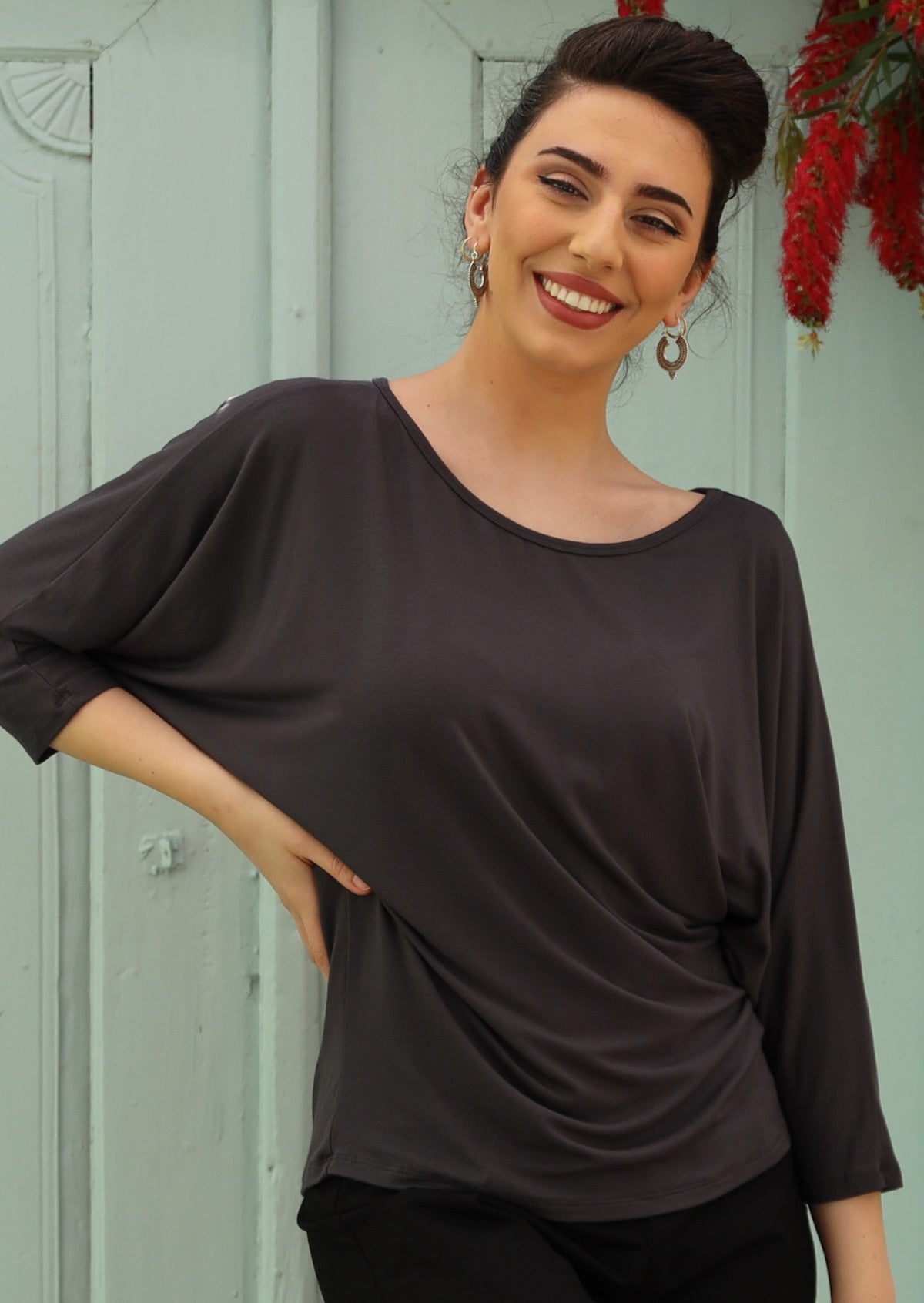 Woman wearing a 3/4 sleeve rayon batwing round neckline dark grey top in front of grey wall.