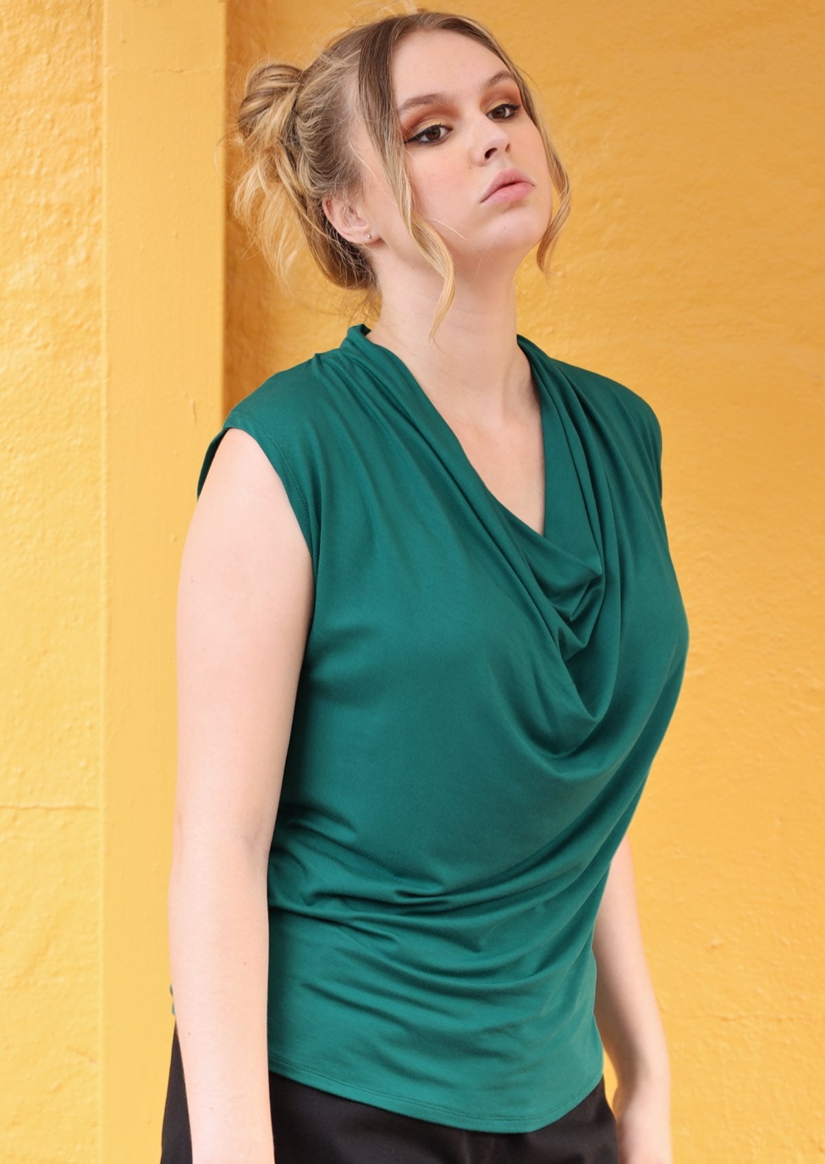 Woman wearing a sleeveless cowl neck jade green top in front of a yellow wall.