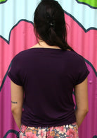 Back view of a woman wearing a wide neck mod purple stretch rayon boat neck top