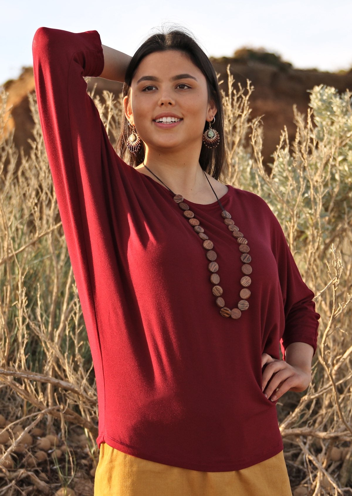 Woman wearing a 3/4 sleeve rayon batwing round neckline maroon top with wooden beads.