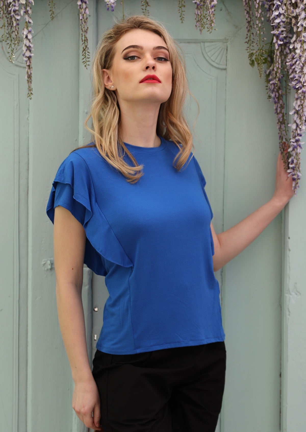 Woman standing in front of grey door wearing a ruffle electric blue round neck short cap sleeve rayon top.